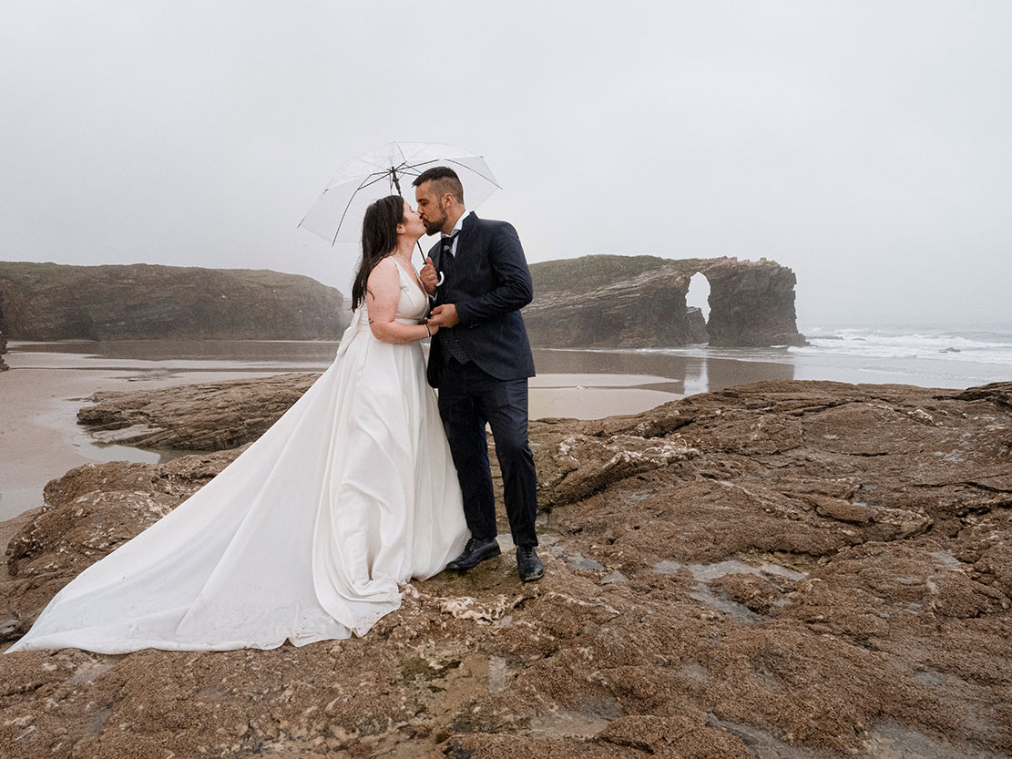 Pareja de novios en postboda en Playa de las Catedrales, Ribadeo, con paraguas en día nublado