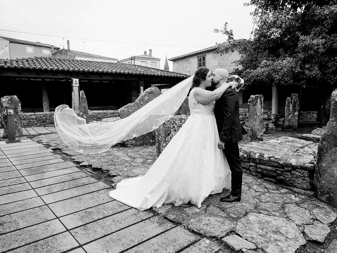 Pareja de novios besándose en una boda rural en Melide, Galicia, con velo al viento – Sanoguera Fotografía